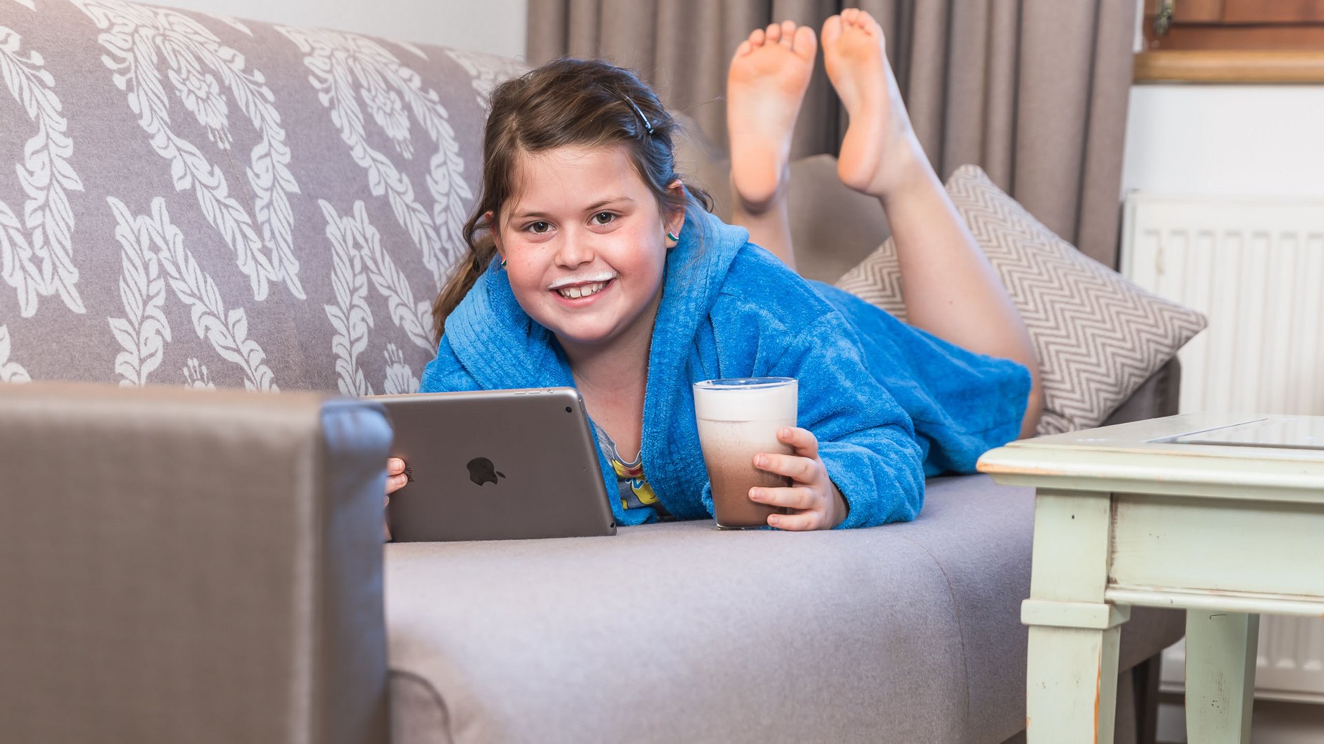 Girl in blue bathrobe with milk mustache drinking milk and holding tablet on sofa