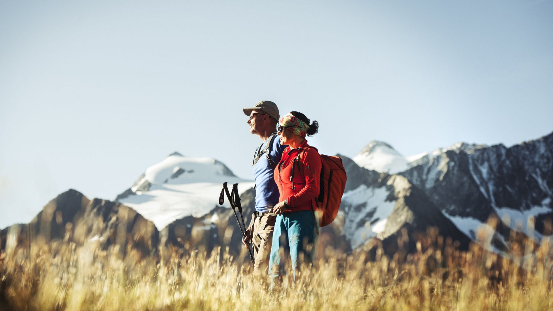Two hikers with backpacks standing on a mountain meadow with snowy peaks in the background