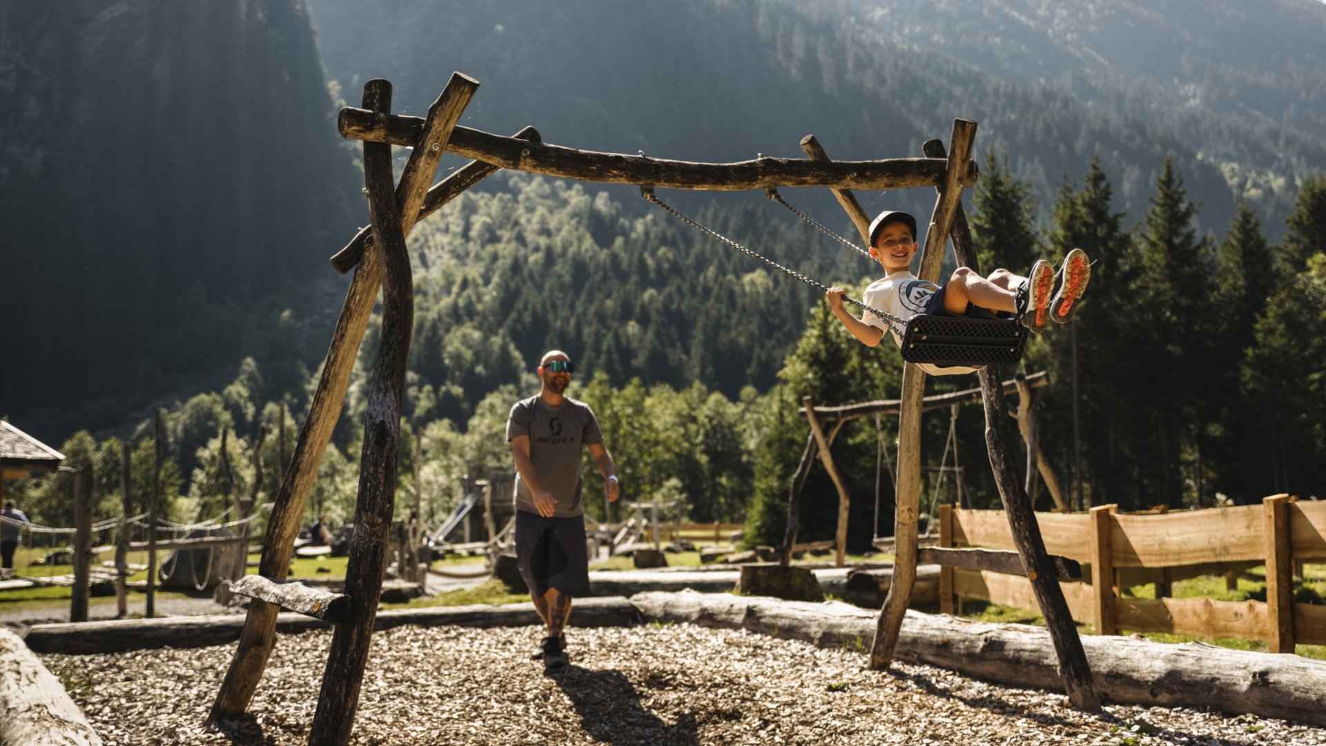 Your family hotel in Neustift im Stubaital Boy swinging pushed by adult on a playground surrounded by mountains and trees