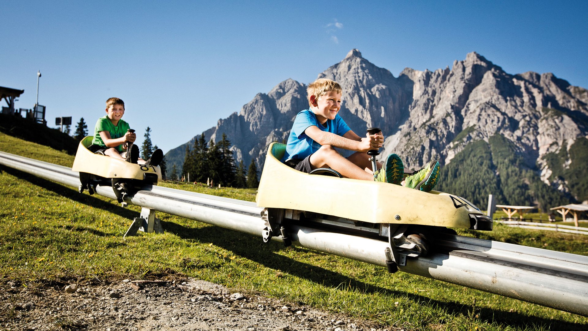 Spannendes rund ums Aktivhotel im Stubaital Zwei Jungen fahren auf Alpenrodelbahn vor Bergkulisse bei Sonnenschein