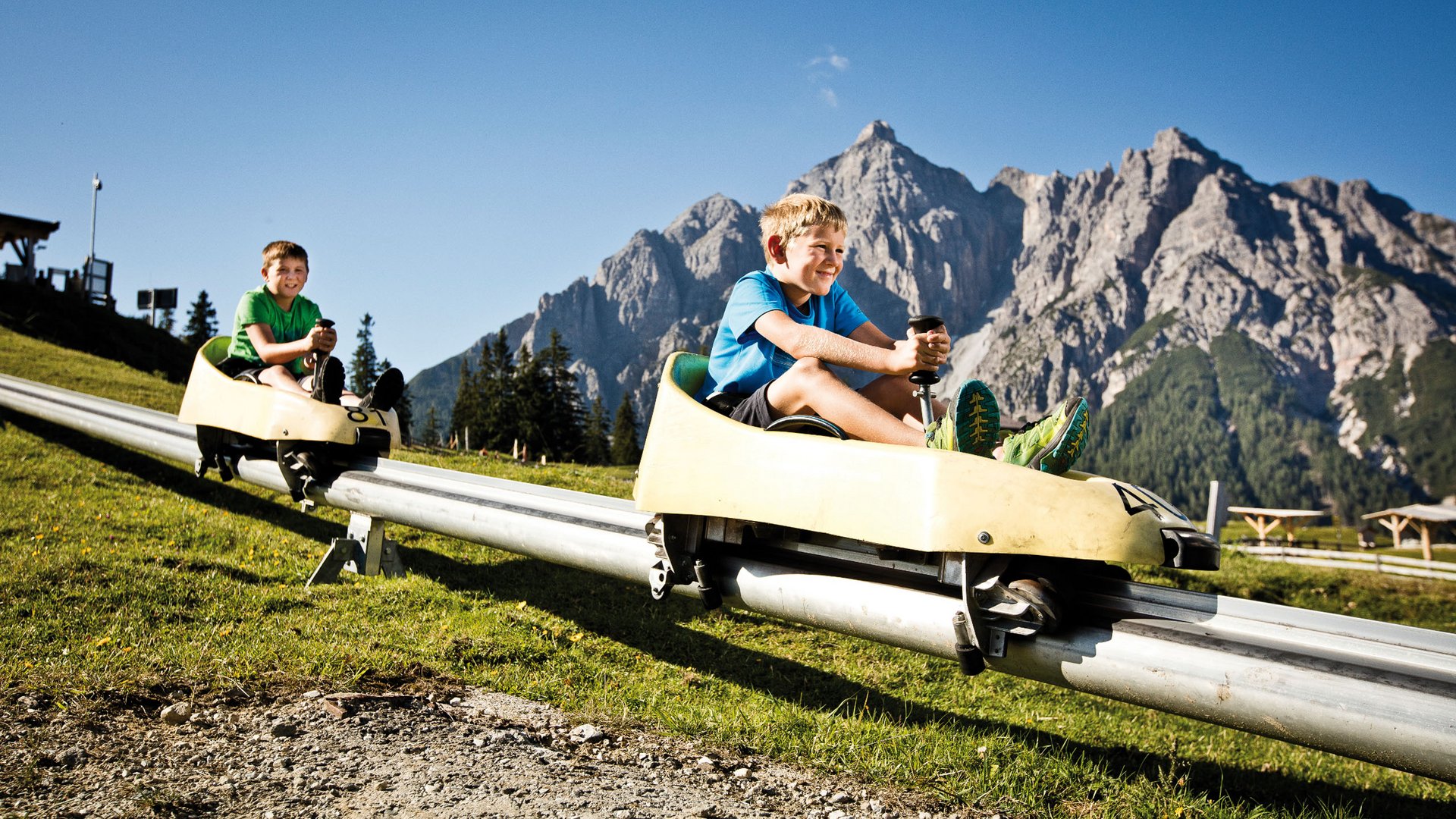 Spannendes rund ums Aktivhotel im Stubaital Zwei Jungen fahren auf Alpenrodelbahn vor Bergkulisse bei Sonnenschein