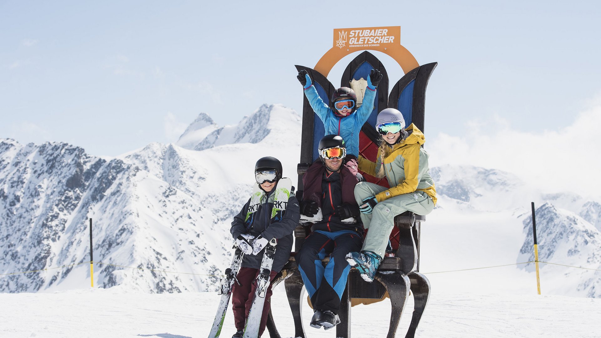 Family with ski helmets sitting on a large chair in front of snowy mountains at Stubai