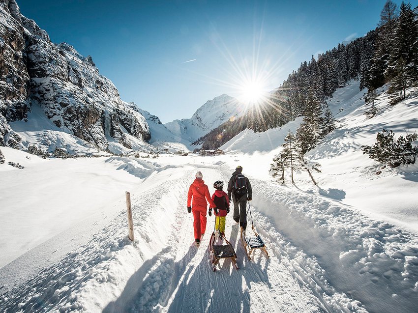 Your family hotel in Neustift im Stubaital Family walking with sleds on snowy mountain trail in bright sunshine