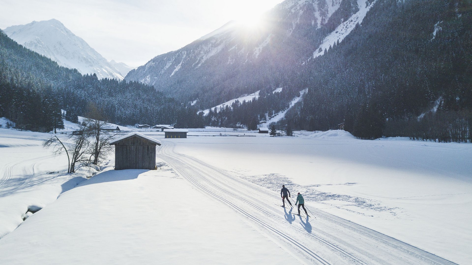 Zwei Langläufer auf verschneiter Spur in einem sonnigen Bergtal mit Hütten