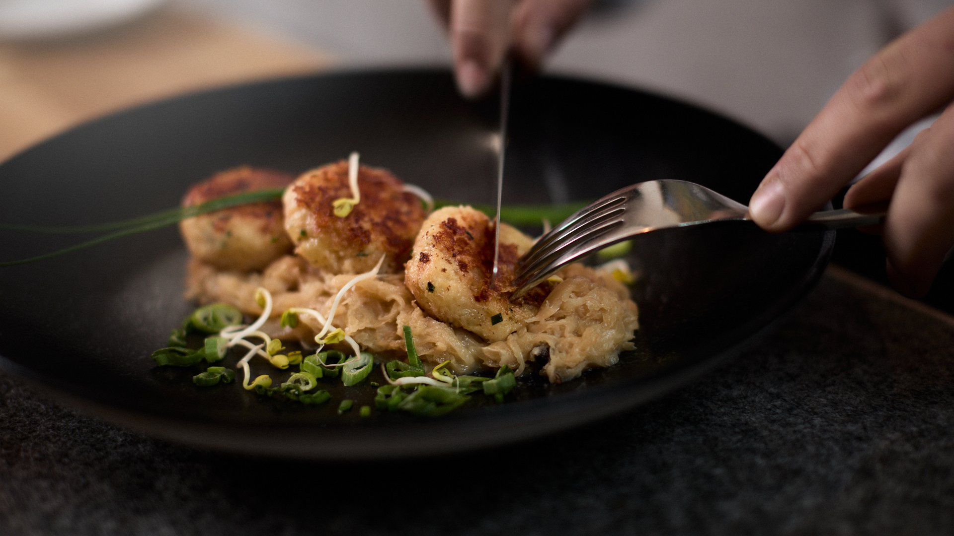 Willkommen im Hotel in Neustift im Stubaital! Person zerteilt gebratene Klöße mit Sauerkraut und Frühlingszwiebeln auf schwarzem Teller